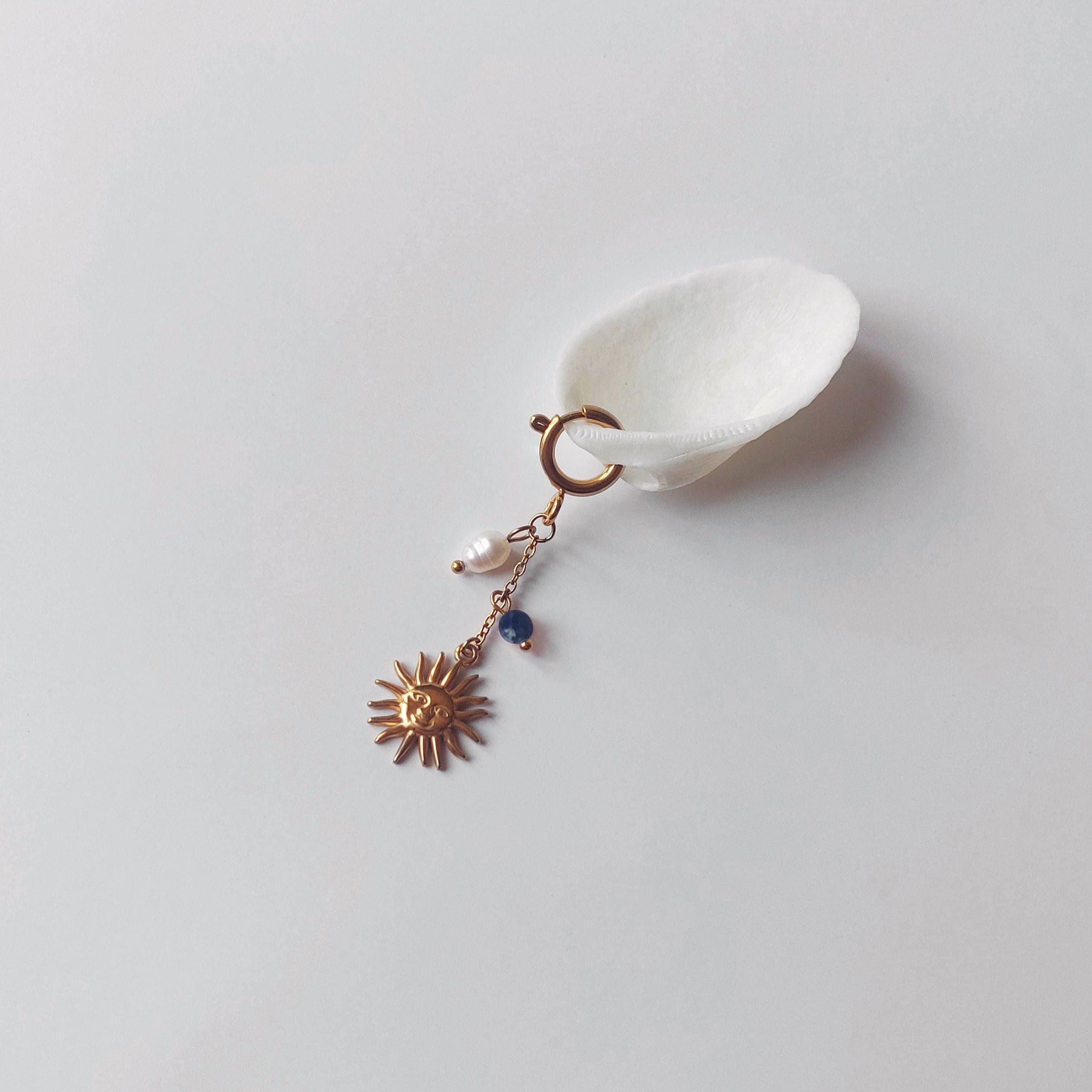 Charm à anneau ressort doré composé d'une perle d'eau douce blanche baroque, d'une chaînette fine ornée d'une petite perle de sodalite et d'un pendentif soleil doré à visage gravé avec rayons, posé contre un coquillage nacré blanc sur fond gris clair.