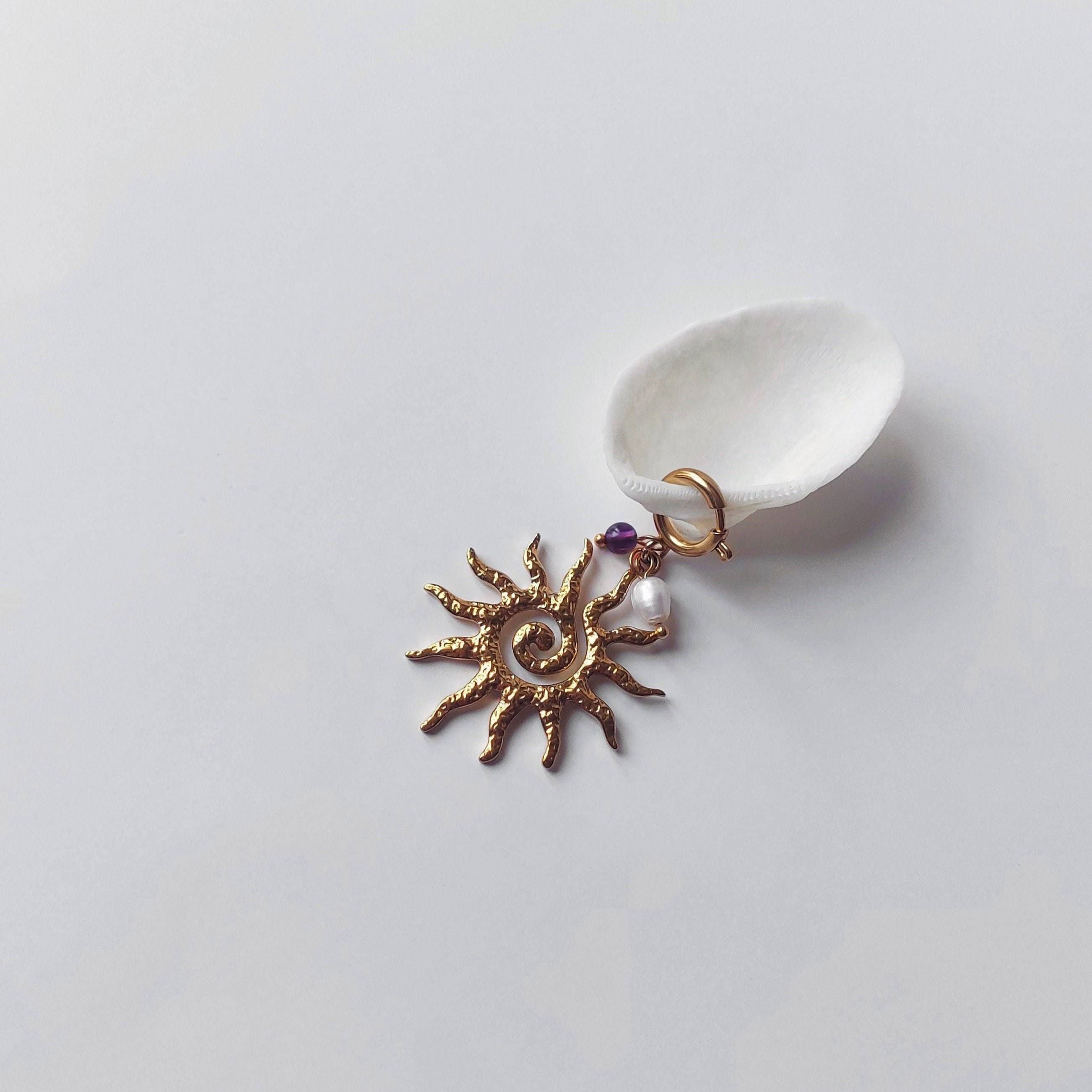 Charm à anneau ressort doré composé d'une petite perle d'améthyste, d'une perle d'eau douce blanche baroque et d'un grand pendentif soleil doré texturé aux rayons irréguliers avec centre en spirale, posé contre un coquillage nacré blanc sur fond gris clair.
