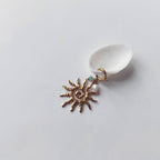Charm à anneau ressort doré composé d'une petite perle d'aventurine, d'une perle d'eau douce blanche baroque et d'un grand pendentif soleil doré texturé aux rayons irréguliers avec centre en spirale, posé contre un coquillage nacré blanc sur fond gris clair.