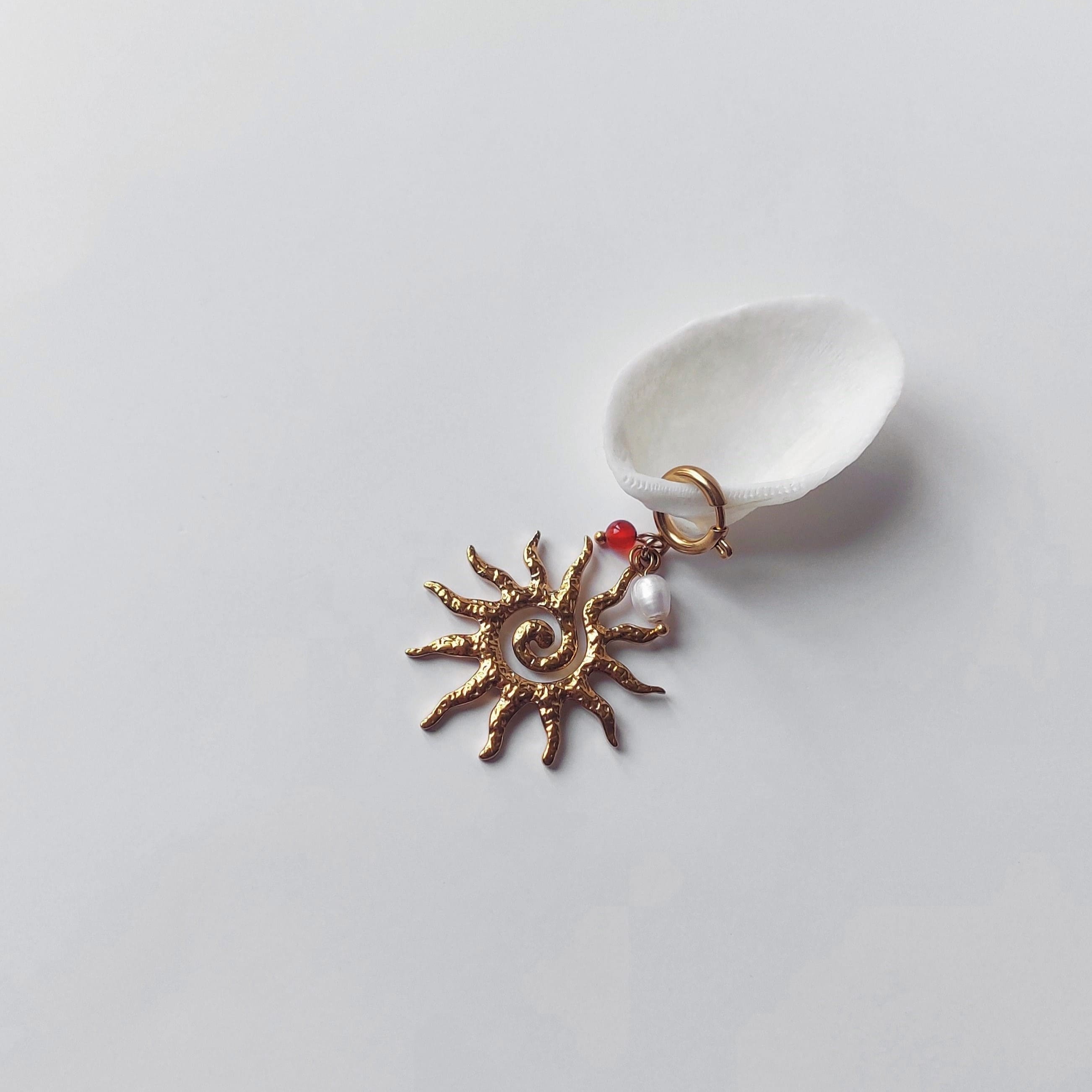Charm à anneau ressort doré composé d'une petite perle de cornaline, d'une perle d'eau douce blanche baroque et d'un grand pendentif soleil doré texturé aux rayons irréguliers avec centre en spirale, posé contre un coquillage nacré blanc sur fond gris clair.