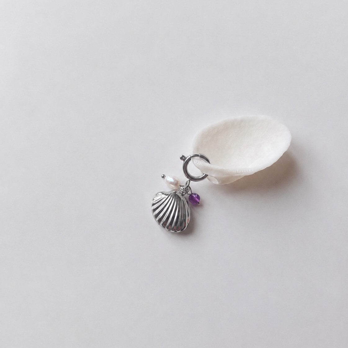 Charm à anneau ressort argenté composé d'une perle d'eau douce blanche, d'une petite perle d'améthyste et d'un pendentif coquille Saint-Jacques argentée striée, posé contre un coquillage nacré blanc sur fond gris clair.