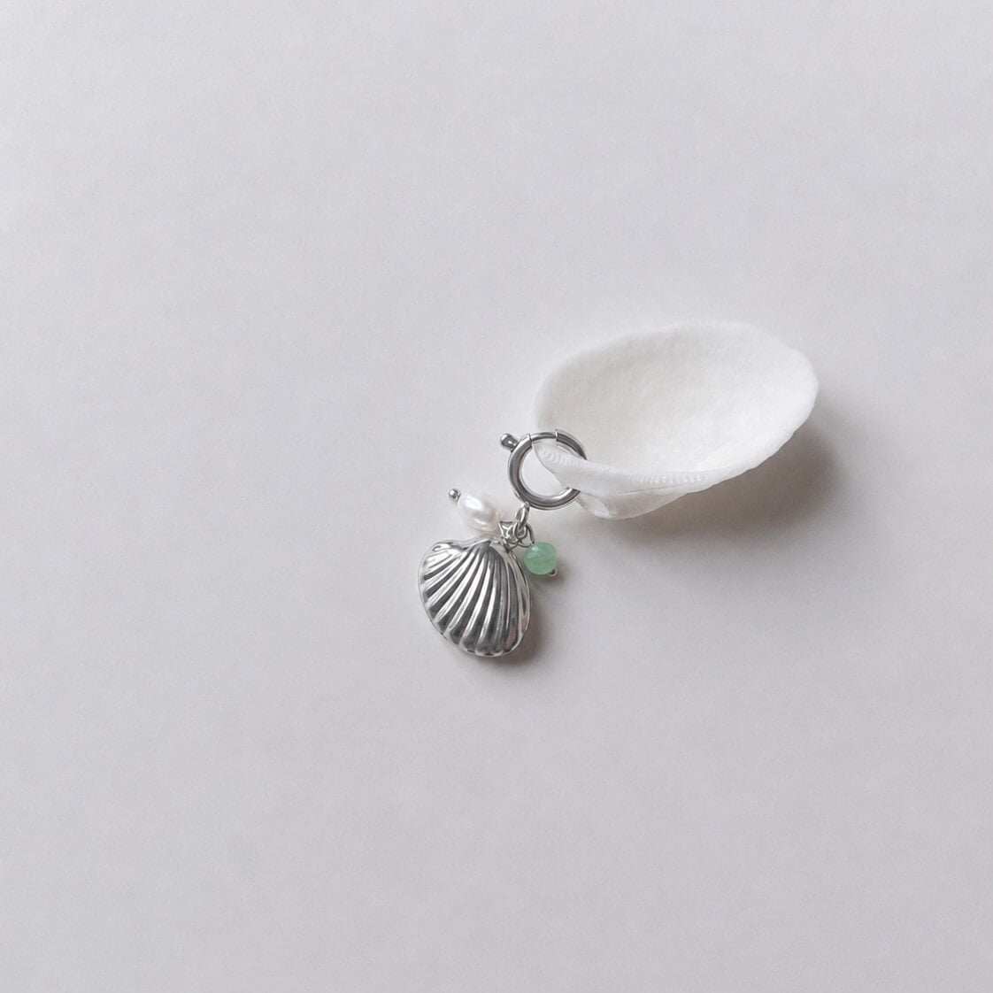 Charm à anneau ressort argenté composé d'une perle d'eau douce blanche, d'une petite perle d'aventurine et d'un pendentif coquille Saint-Jacques argentée striée, posé contre un coquillage nacré blanc sur fond gris clair.