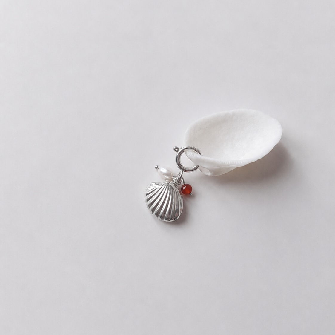 Charm à anneau ressort argenté composé d'une perle d'eau douce blanche, d'une petite perle de cornaline et d'un pendentif coquille Saint-Jacques argentée striée, posé contre un coquillage nacré blanc sur fond gris clair.