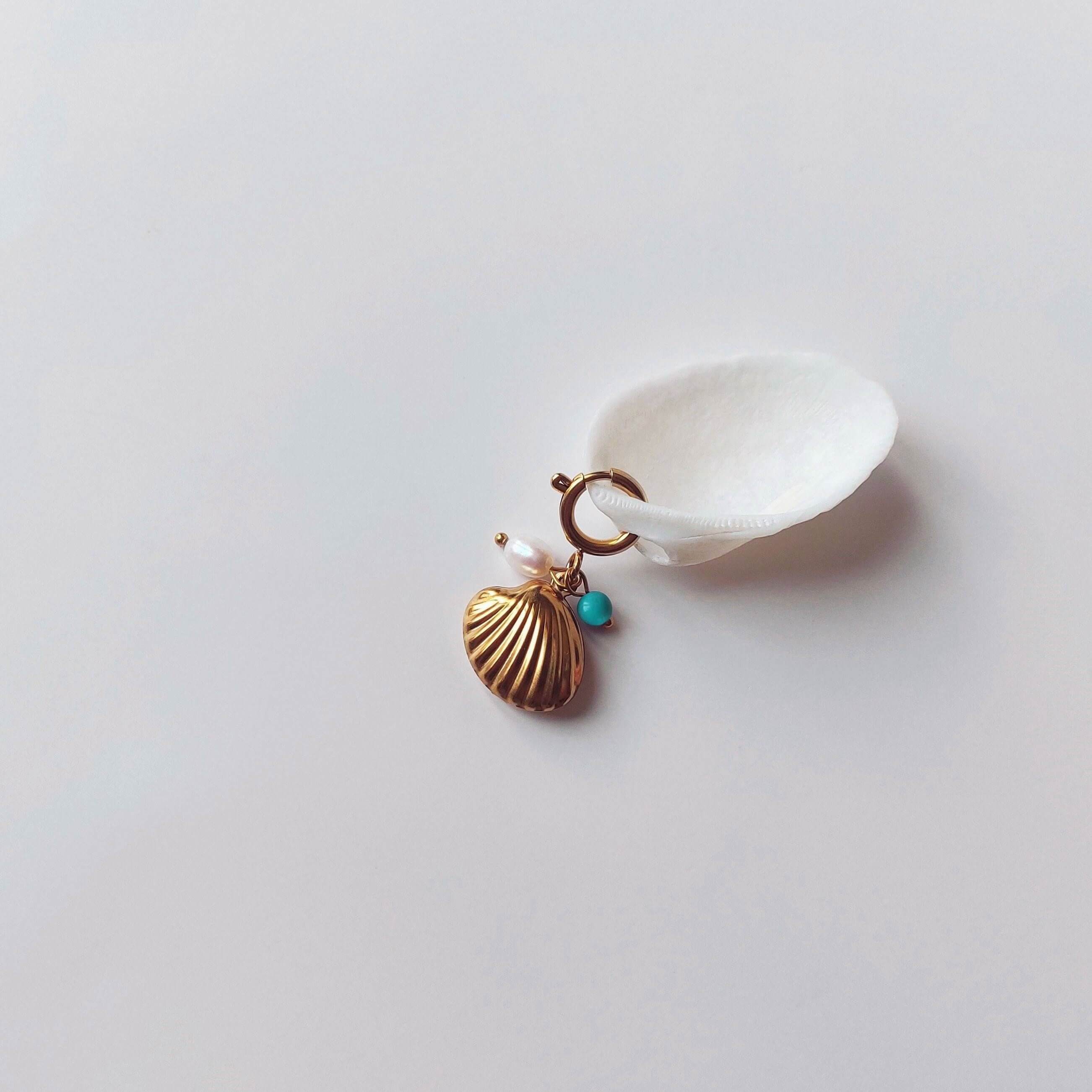 Charm à anneau ressort doré composé d'une perle d'eau douce blanche, d'une petite perle de turquoise et d'un pendentif coquille Saint-Jacques dorée striée, posé contre un coquillage nacré blanc sur fond gris clair.
