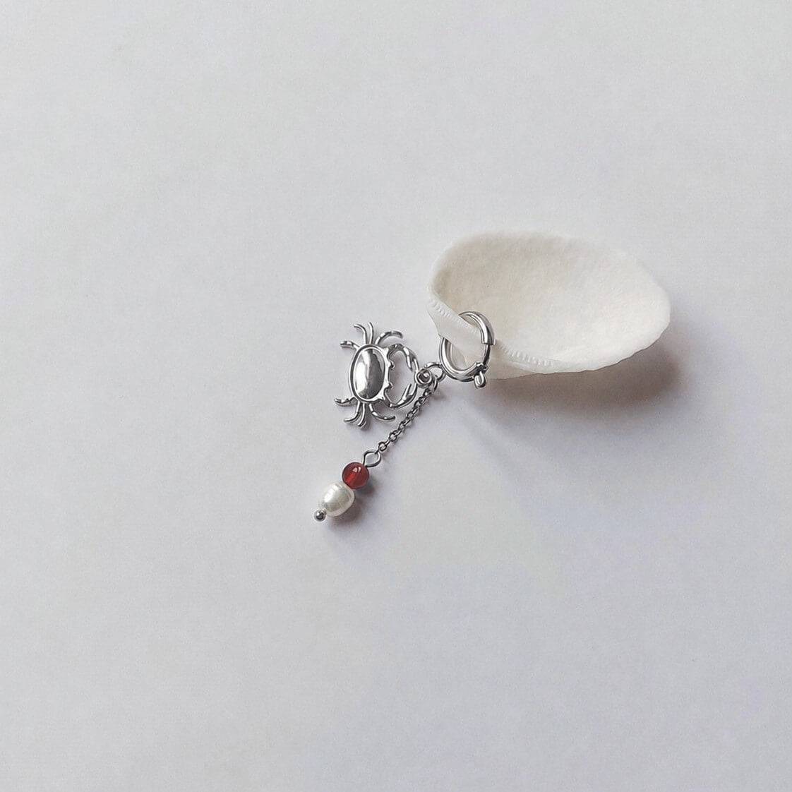 Charm créole argentée en forme de crabe stylisé avec une chaînette fine ornée d'une perle de pierre en cornaline et d'une perle d'eau douce blanche baroque en pendentif, posé contre un coquillage nacré blanc sur fond gris clair.