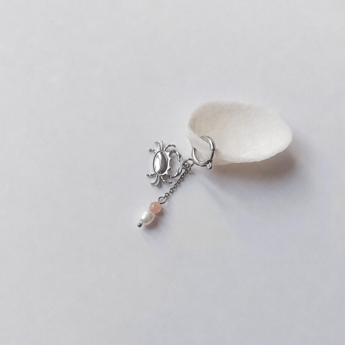 Charm créole argentée en forme de crabe stylisé avec une chaînette fine ornée d'une perle de pierre en pierre de soleil et d'une perle d'eau douce blanche baroque en pendentif, posé contre un coquillage nacré blanc sur fond gris clair.