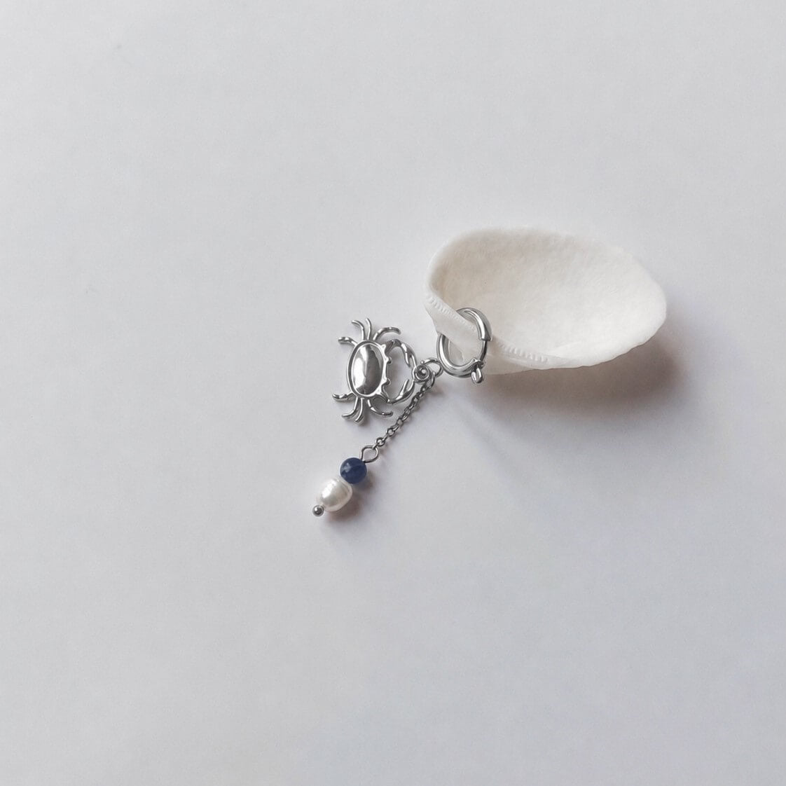 Charm créole argentée en forme de crabe stylisé avec une chaînette fine ornée d'une perle de pierre en sodalite et d'une perle d'eau douce blanche baroque en pendentif, posé contre un coquillage nacré blanc sur fond gris clair.