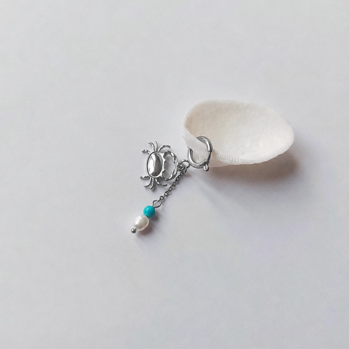Charm créole argentée en forme de crabe stylisé avec une chaînette fine ornée d'une perle de pierre en turquoise et d'une perle d'eau douce blanche baroque en pendentif, posé contre un coquillage nacré blanc sur fond gris clair.