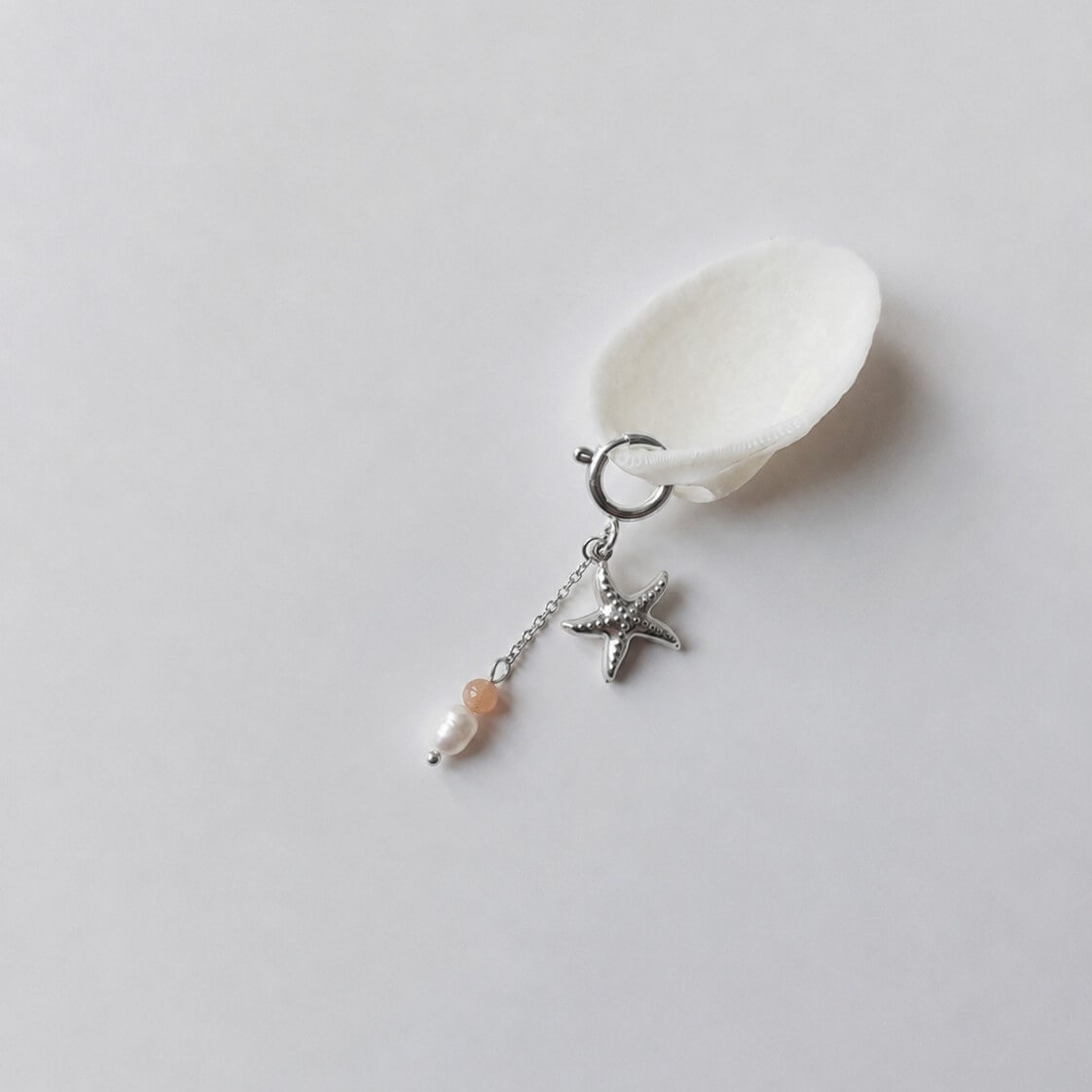 Charm à anneau ressort argenté composé d'un pendentif étoile de mer argentée texturée et d'une chaînette fine ornée d'une petite perle de pierre de soleil et d'une perle d'eau douce blanche baroque, posé contre un coquillage nacré blanc sur fond gris clair.