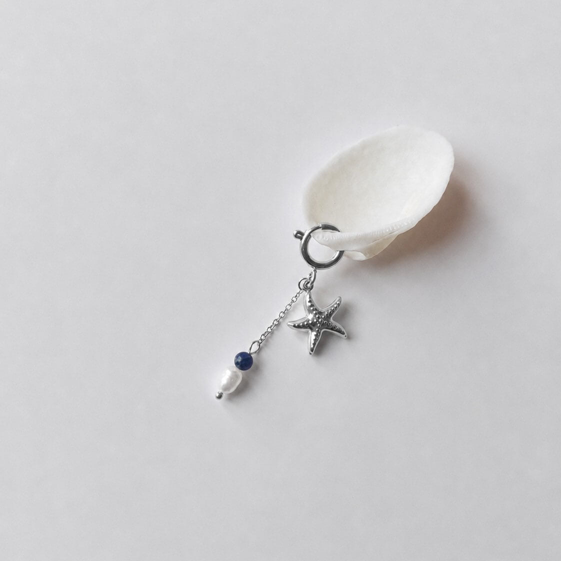 Charm à anneau ressort argenté composé d'un pendentif étoile de mer argentée texturée et d'une chaînette fine ornée d'une petite perle de sodalite et d'une perle d'eau douce blanche baroque, posé contre un coquillage nacré blanc sur fond gris clair.