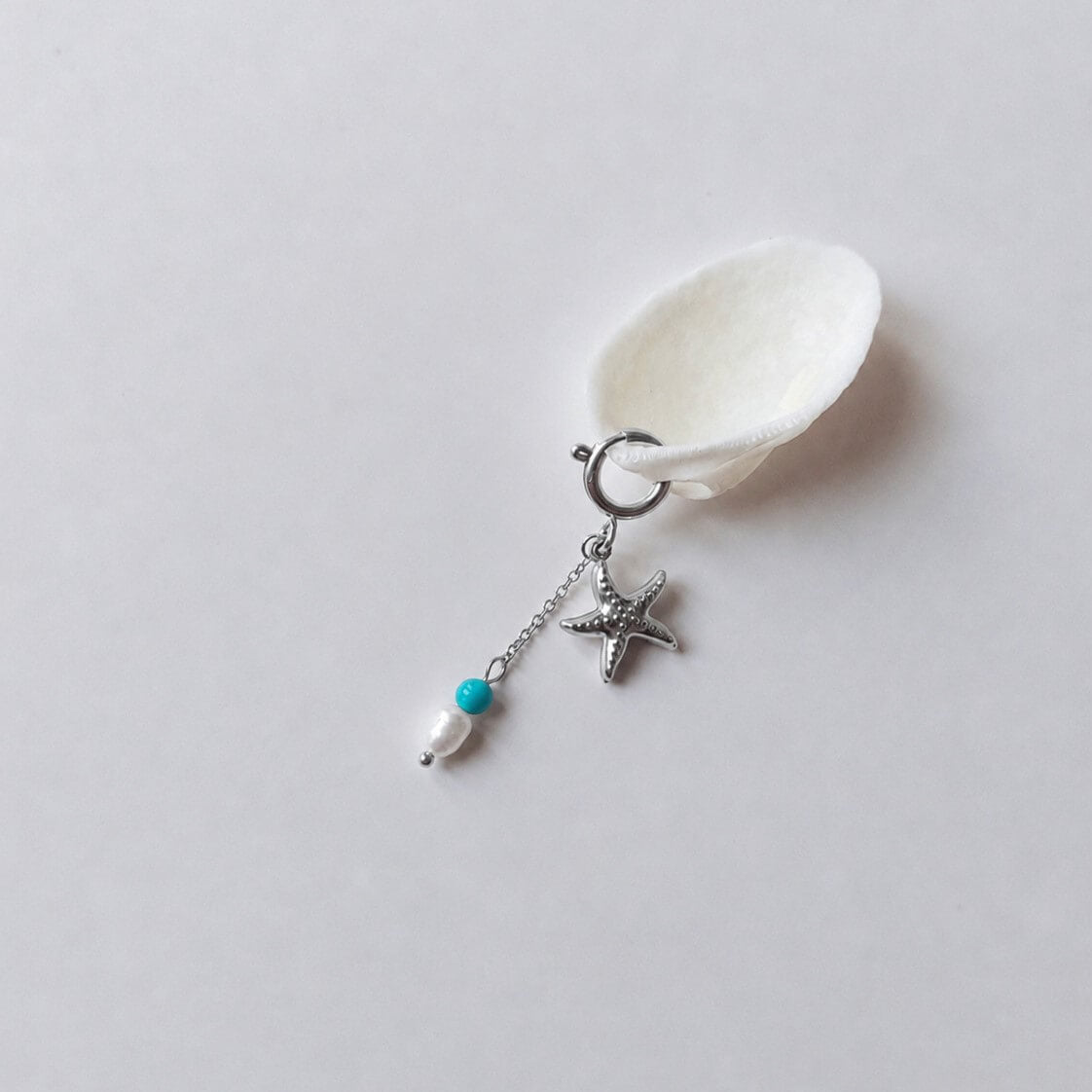 Charm à anneau ressort argenté composé d'un pendentif étoile de mer argentée texturée et d'une chaînette fine ornée d'une petite perle de turquoise et d'une perle d'eau douce blanche baroque, posé contre un coquillage nacré blanc sur fond gris clair.