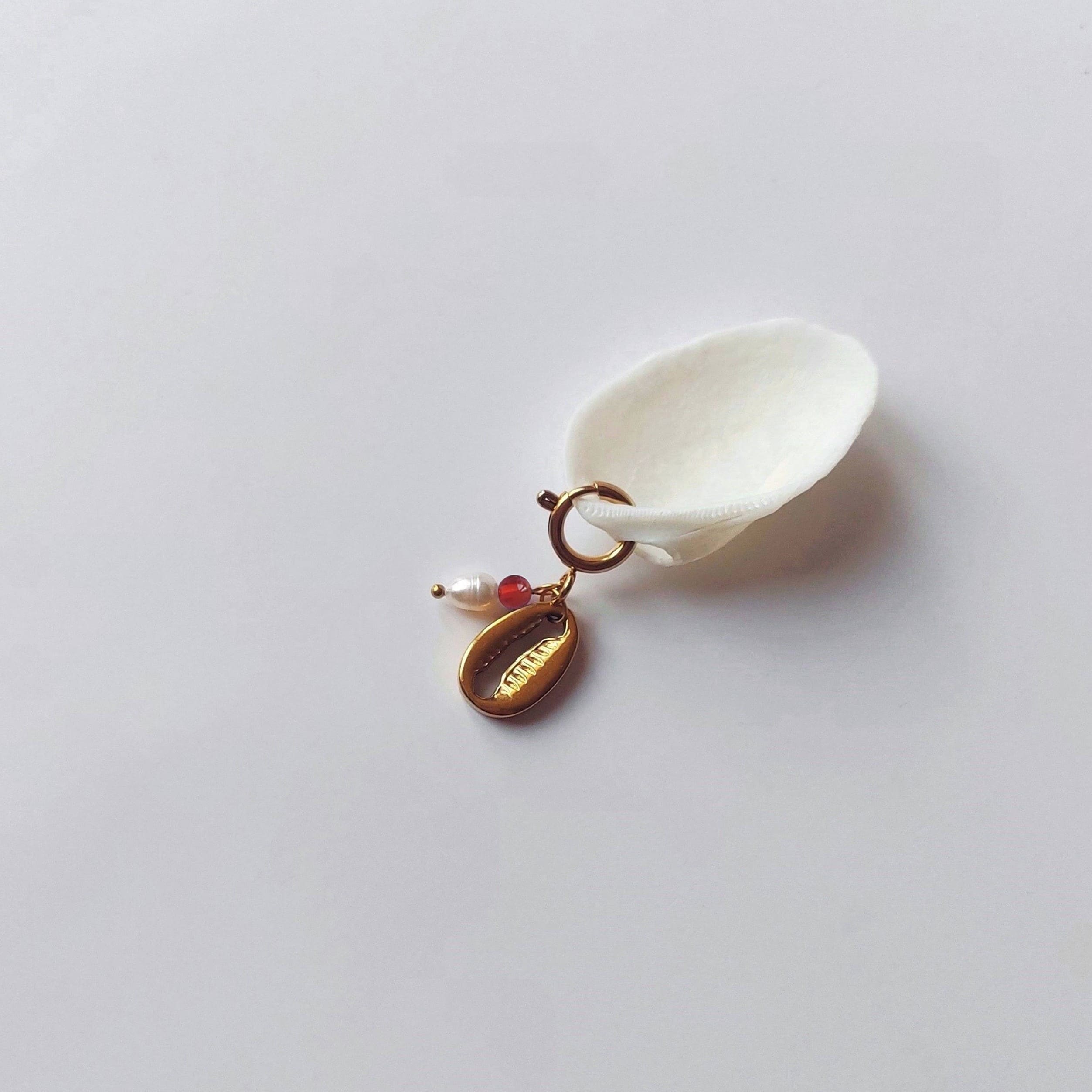 Charm à anneau ressort doré composé d'une perle d'eau douce blanche, d'une petite perle de cornalinel et d'un pendentif cauri doré, posé contre un coquillage nacré blanc sur fond gris clair.