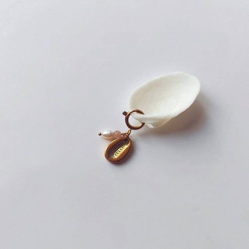 Charm à anneau ressort doré composé d'une perle d'eau douce blanche, d'une petite perle de pierre de soleil et d'un pendentif cauri doré, posé contre un coquillage nacré blanc sur fond gris clair.