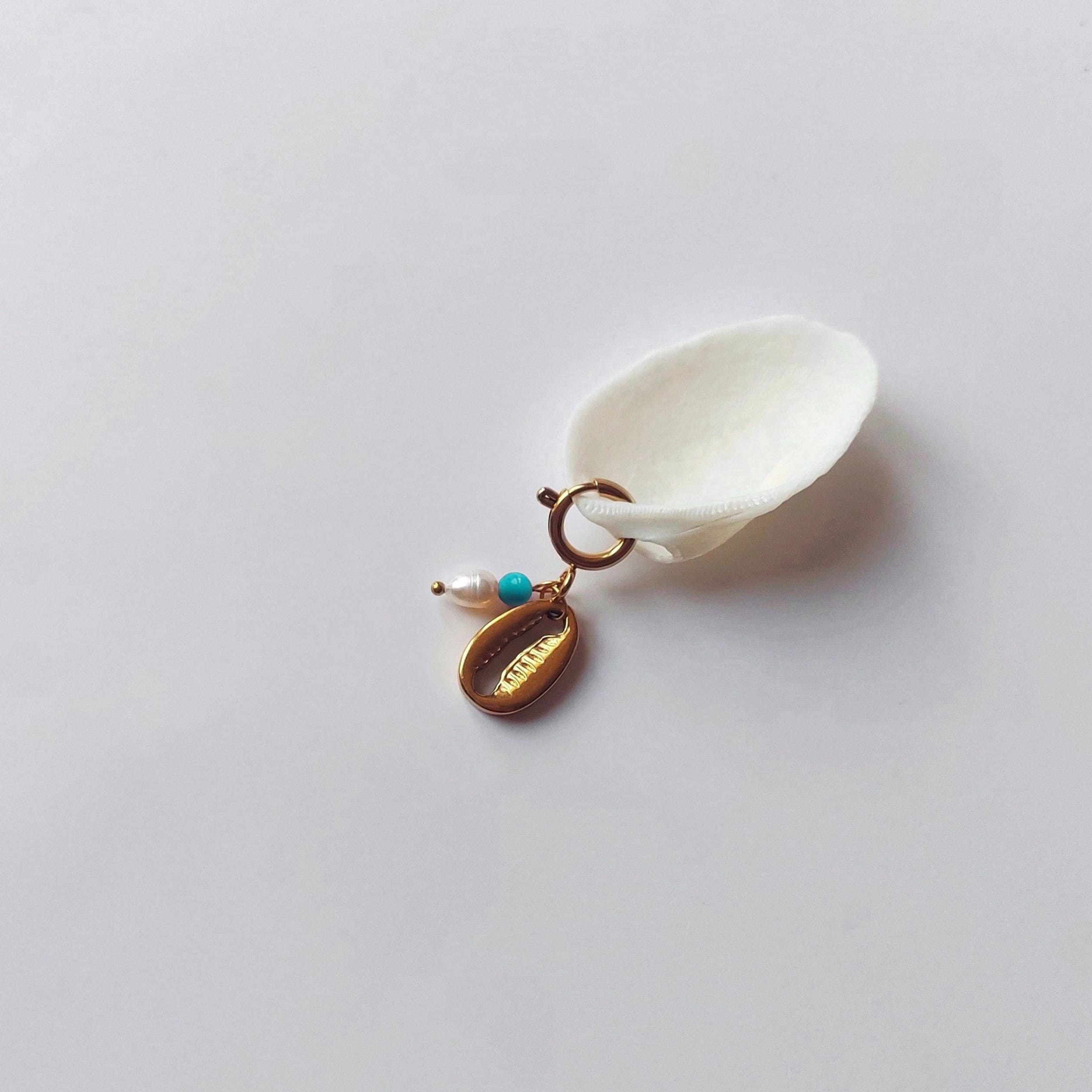 Charm à anneau ressort doré composé d'une perle d'eau douce blanche, d'une petite perle de turquoise et d'un pendentif cauri doré, posé contre un coquillage nacré blanc sur fond gris clair.