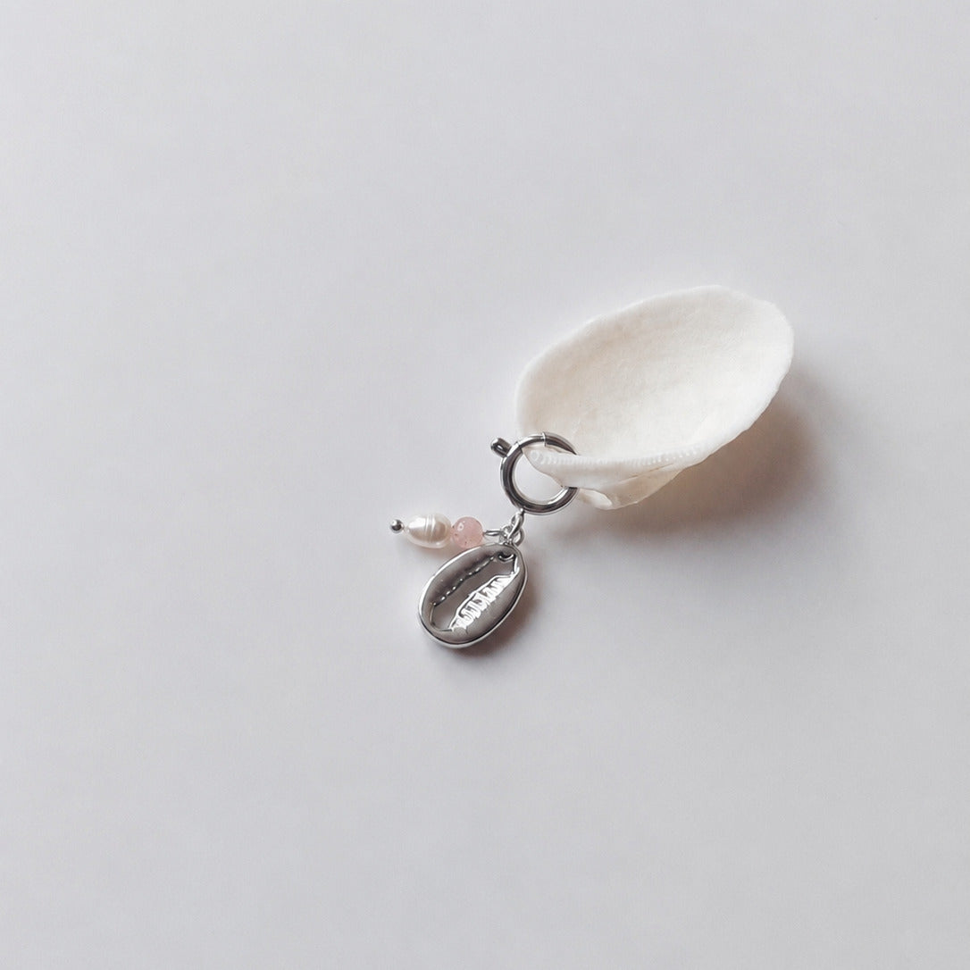 Charm à anneau ressort argenté composé d'une perle d'eau douce blanche, d'une petite perle de pierre de soleil et d'un pendentif cauri argenté, posé contre un coquillage nacré blanc sur fond gris clair.
