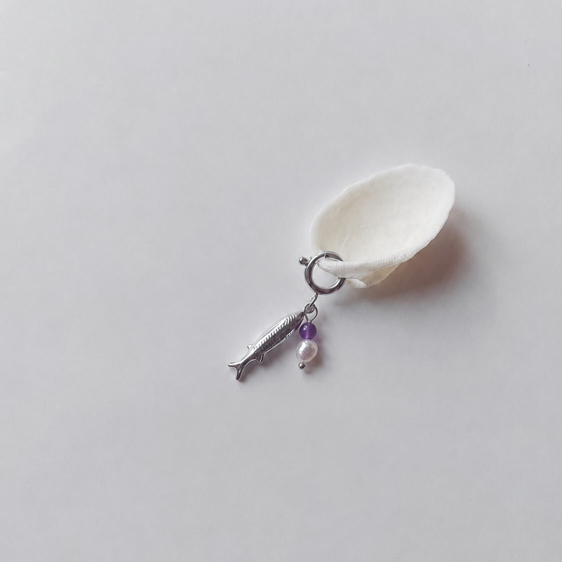 Charm à anneau ressort argenté composé d'un pendentif poisson argenté gravé, d'une petite perle d'améthyste et d'une perle d'eau douce blanche baroque, posé contre un coquillage nacré blanc sur fond gris clair.