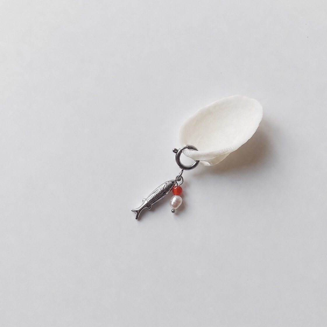 Charm à anneau ressort argenté composé d'un pendentif poisson argenté gravé, d'une petite perle de cornaline et d'une perle d'eau douce blanche baroque, posé contre un coquillage nacré blanc sur fond gris clair.