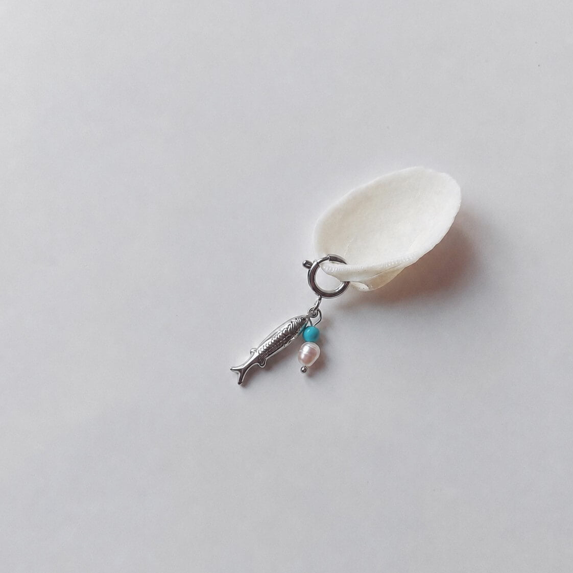 Charm à anneau ressort argenté composé d'un pendentif poisson argenté gravé, d'une petite perle de turquoise et d'une perle d'eau douce blanche baroque, posé contre un coquillage nacré blanc sur fond gris clair.
