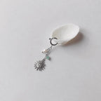 Charm à anneau ressort argenté composé d'une perle d'eau douce blanche baroque, d'une chaînette fine ornée d'une petite perle d'aventurine verte et d'un pendentif soleil argenté à visage gravé avec rayons, posé contre un coquillage nacré blanc sur fond gris clair.