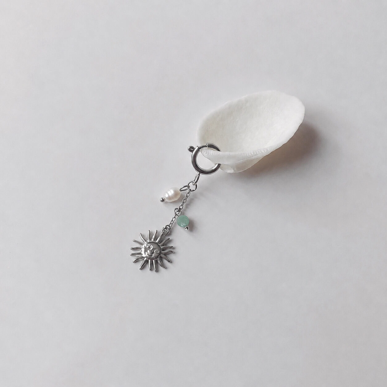 Charm à anneau ressort argenté composé d'une perle d'eau douce blanche baroque, d'une chaînette fine ornée d'une petite perle d'aventurine verte et d'un pendentif soleil argenté à visage gravé avec rayons, posé contre un coquillage nacré blanc sur fond gris clair.