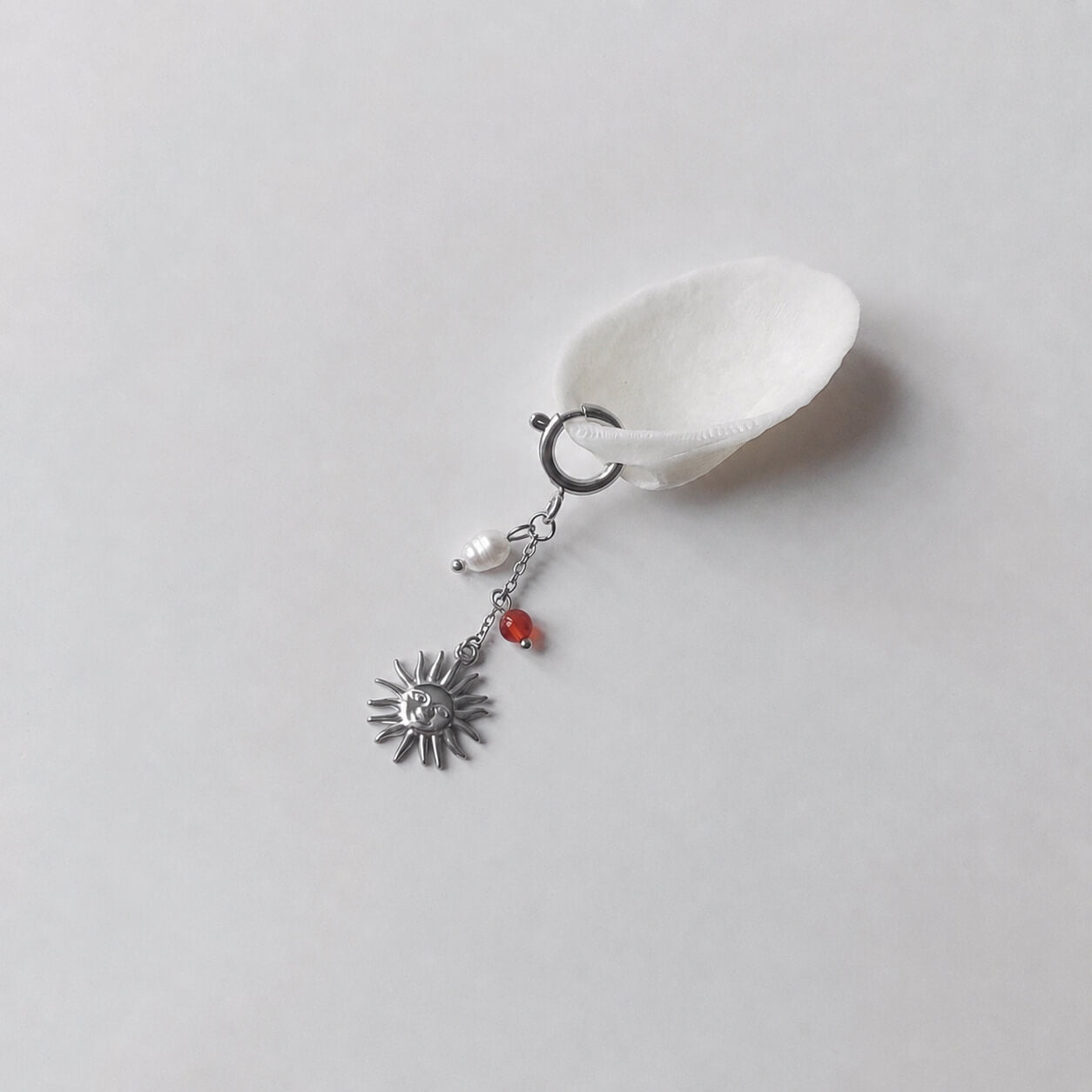 Charm à anneau ressort argenté composé d'une perle d'eau douce blanche baroque, d'une chaînette fine ornée d'une petite perle de cornaline et d'un pendentif soleil argenté à visage gravé avec rayons, posé contre un coquillage nacré blanc sur fond gris clair.
