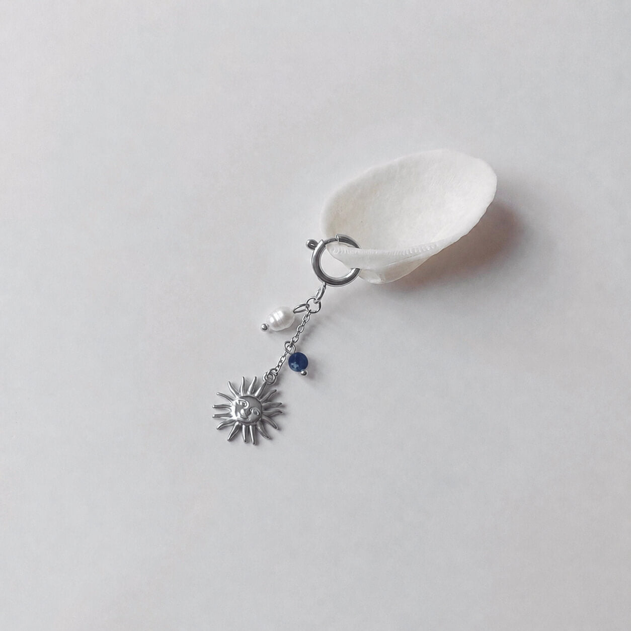 Charm à anneau ressort argenté composé d'une perle d'eau douce blanche baroque, d'une chaînette fine ornée d'une petite perle de sodalite et d'un pendentif soleil argenté à visage gravé avec rayons, posé contre un coquillage nacré blanc sur fond gris clair.