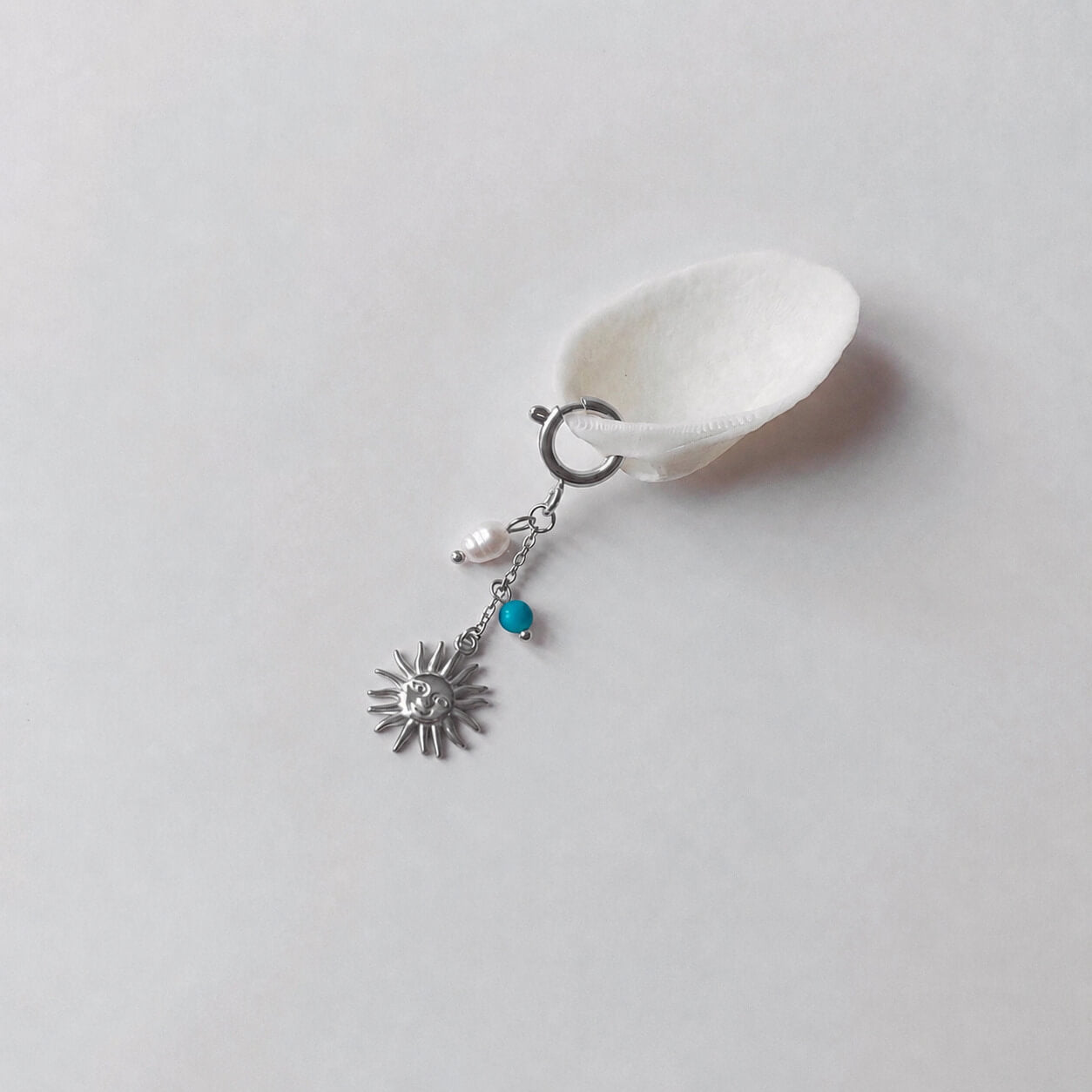 Charm à anneau ressort argenté composé d'une perle d'eau douce blanche baroque, d'une chaînette fine ornée d'une petite perle de turquoise et d'un pendentif soleil argenté à visage gravé avec rayons, posé contre un coquillage nacré blanc sur fond gris clair.