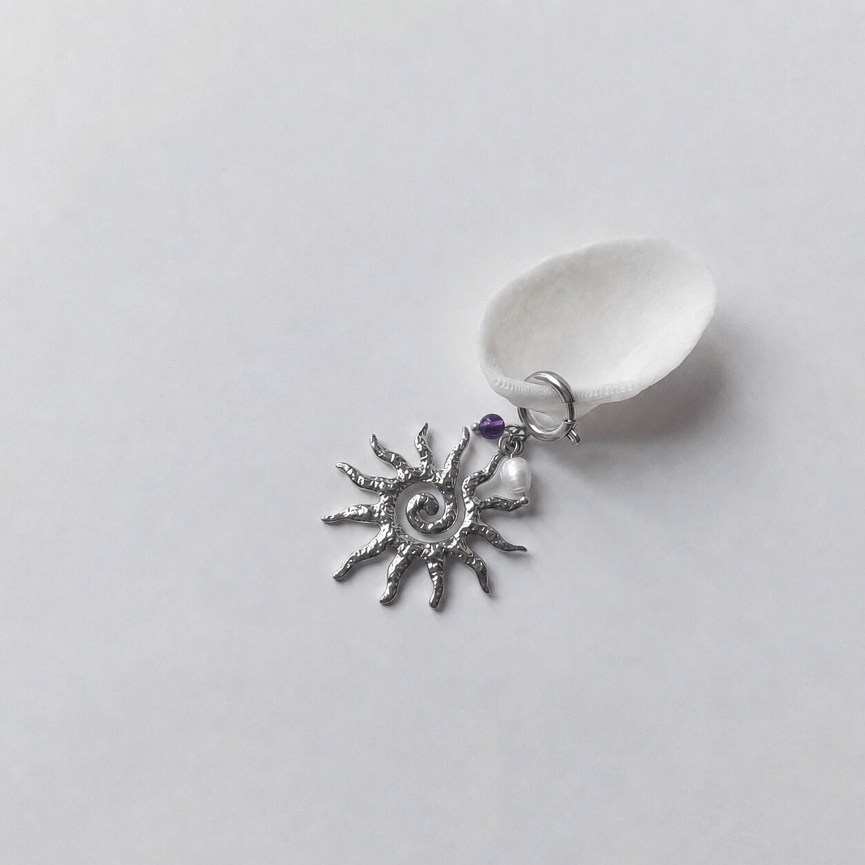 Charm à anneau ressort argenté composé d'une petite perle d'améthyste, d'une perle d'eau douce blanche baroque et d'un grand pendentif soleil argenté texturé aux rayons irréguliers avec centre en spirale, posé contre un coquillage nacré blanc sur fond gris clair.