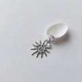 Charm à anneau ressort argenté composé d'une petite perle d'aventurine, d'une perle d'eau douce blanche baroque et d'un grand pendentif soleil argenté texturé aux rayons irréguliers avec centre en spirale, posé contre un coquillage nacré blanc sur fond gris clair.