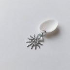 Charm à anneau ressort argenté composé d'une petite perle d'aventurine, d'une perle d'eau douce blanche baroque et d'un grand pendentif soleil argenté texturé aux rayons irréguliers avec centre en spirale, posé contre un coquillage nacré blanc sur fond gris clair.