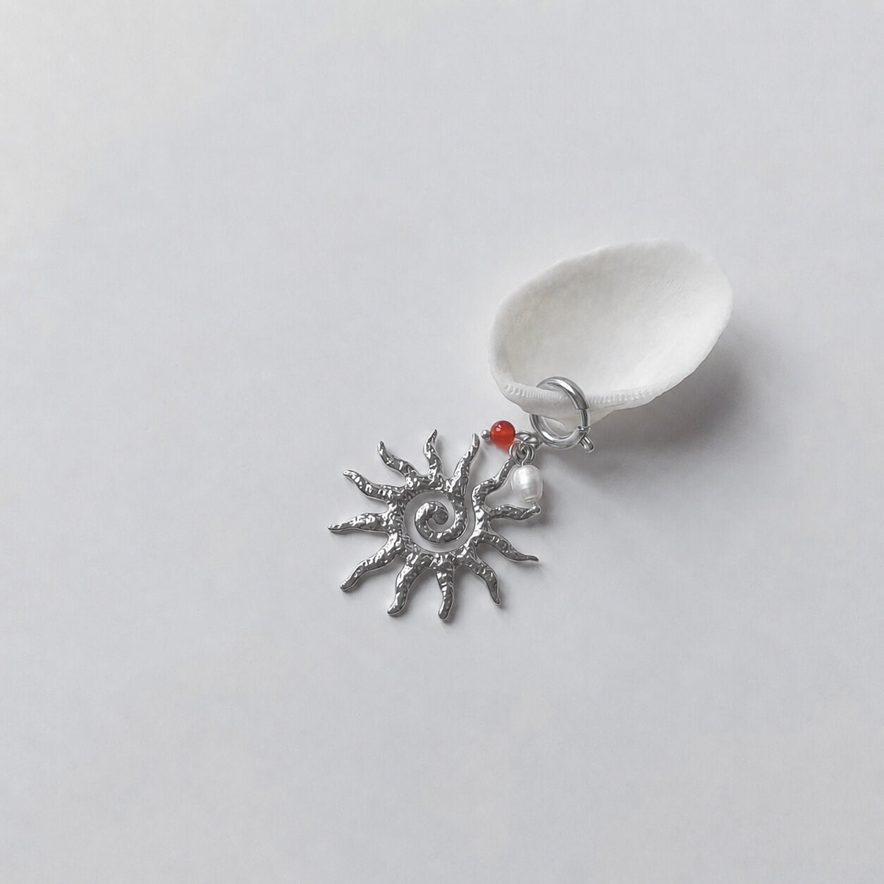 Charm à anneau ressort argenté composé d'une petite perle de cornaline, d'une perle d'eau douce blanche baroque et d'un grand pendentif soleil argenté texturé aux rayons irréguliers avec centre en spirale, posé contre un coquillage nacré blanc sur fond gris clair.