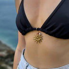 Charm grand soleil doré texturé aux rayons irréguliers avec centre en spirale, accompagné d'une petite perle de cornaline rouge et d'une perle d'eau douce blanche baroque en pendentif, attaché à l'anneau central d'un haut de bikini triangle noir, porté par une femme en jean avec un fond de mer et ciel bleu en bokeh.