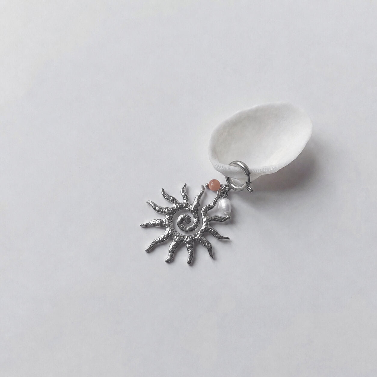 Charm à anneau ressort argenté composé d'une petite perle de pierre de soleil, d'une perle d'eau douce blanche baroque et d'un grand pendentif soleil argenté texturé aux rayons irréguliers avec centre en spirale, posé contre un coquillage nacré blanc sur fond gris clair.