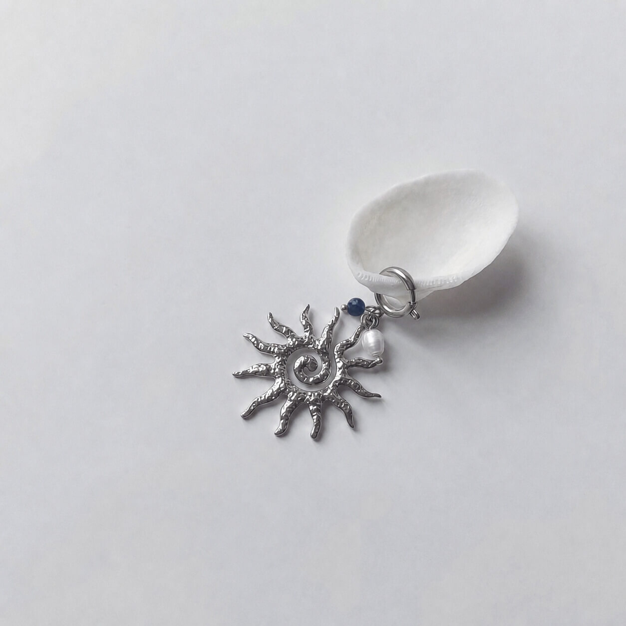 Charm à anneau ressort argenté composé d'une petite perle de sodalite, d'une perle d'eau douce blanche baroque et d'un grand pendentif soleil argenté texturé aux rayons irréguliers avec centre en spirale, posé contre un coquillage nacré blanc sur fond gris clair.