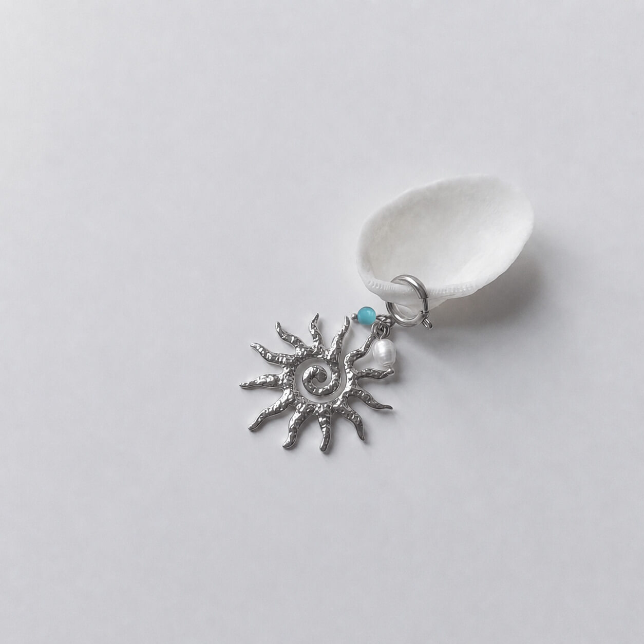 Charm à anneau ressort argenté composé d'une petite perle de turquoise, d'une perle d'eau douce blanche baroque et d'un grand pendentif soleil argenté texturé aux rayons irréguliers avec centre en spirale, posé contre un coquillage nacré blanc sur fond gris clair.