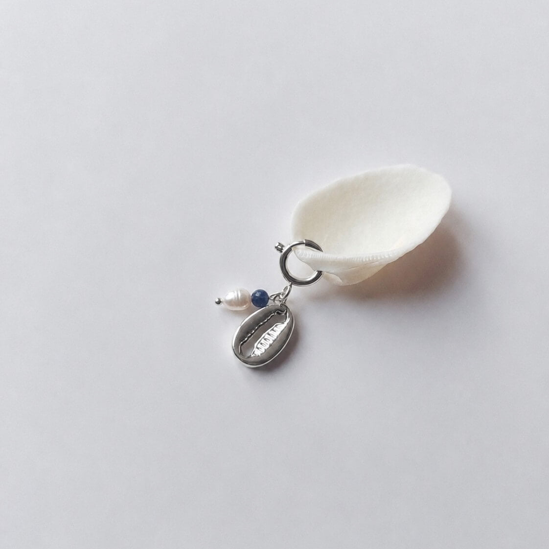 Charm à anneau ressort argenté composé d'une perle d'eau douce blanche, d'une petite perle de sodalitel et d'un pendentif cauri argenté, posé contre un coquillage nacré blanc sur fond gris clair.