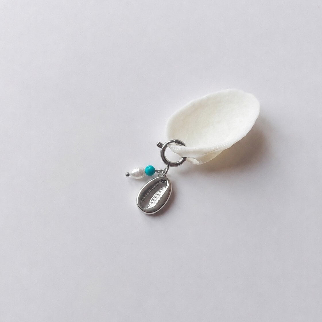 Charm à anneau ressort argenté composé d'une perle d'eau douce blanche, d'une petite perle de turquoise et d'un pendentif cauri argenté, posé contre un coquillage nacré blanc sur fond gris clair.