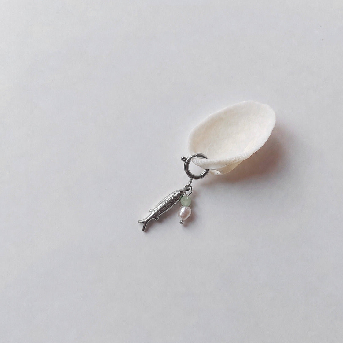Charm à anneau ressort argenté composé d'un pendentif poisson argenté gravé, d'une petite perle d'aventurine et d'une perle d'eau douce blanche baroque, posé contre un coquillage nacré blanc sur fond gris clair.
