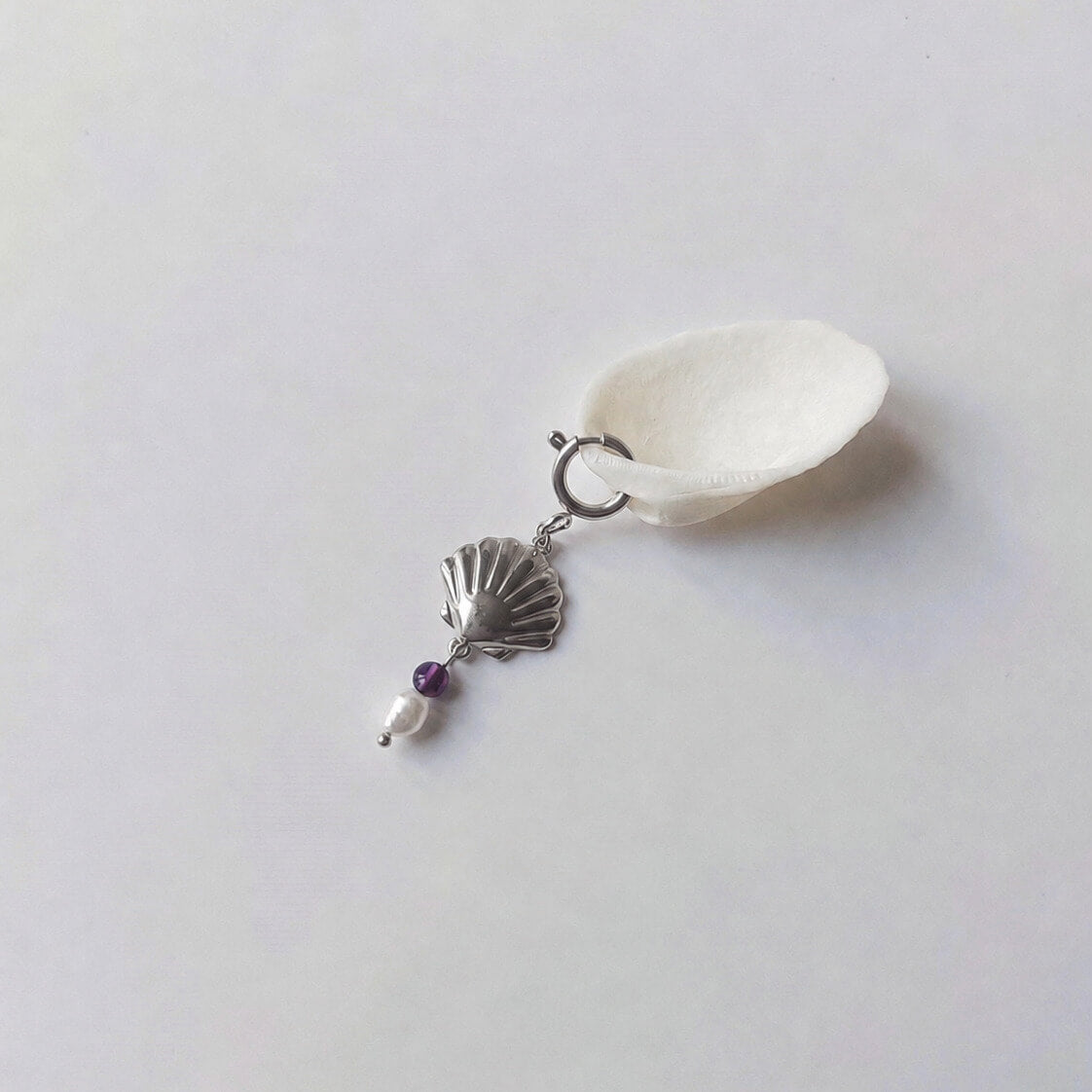 Charm à anneau ressort argenté composé d'un pendentif coquille Saint-Jacques argentée striée, d'une petite perle d'améthyste et d'une perle d'eau douce blanche baroque, posé contre un coquillage nacré blanc sur fond gris clair.