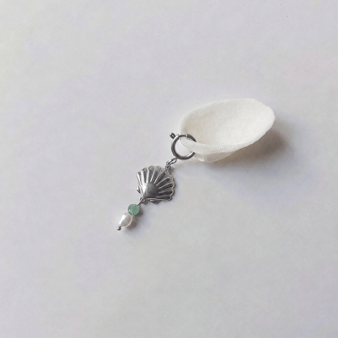 Charm à anneau ressort argenté composé d'un pendentif coquille Saint-Jacques argentée striée, d'une petite perle d'aventurine et d'une perle d'eau douce blanche baroque, posé contre un coquillage nacré blanc sur fond gris clair.