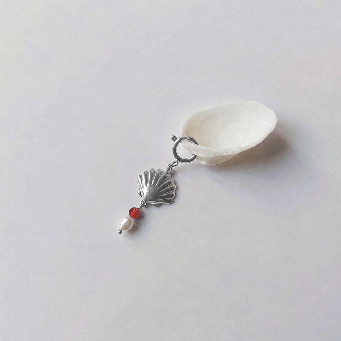 Charm à anneau ressort argenté composé d'un pendentif coquille Saint-Jacques argentée striée, d'une petite perle de cornaline et d'une perle d'eau douce blanche baroque, posé contre un coquillage nacré blanc sur fond gris clair.