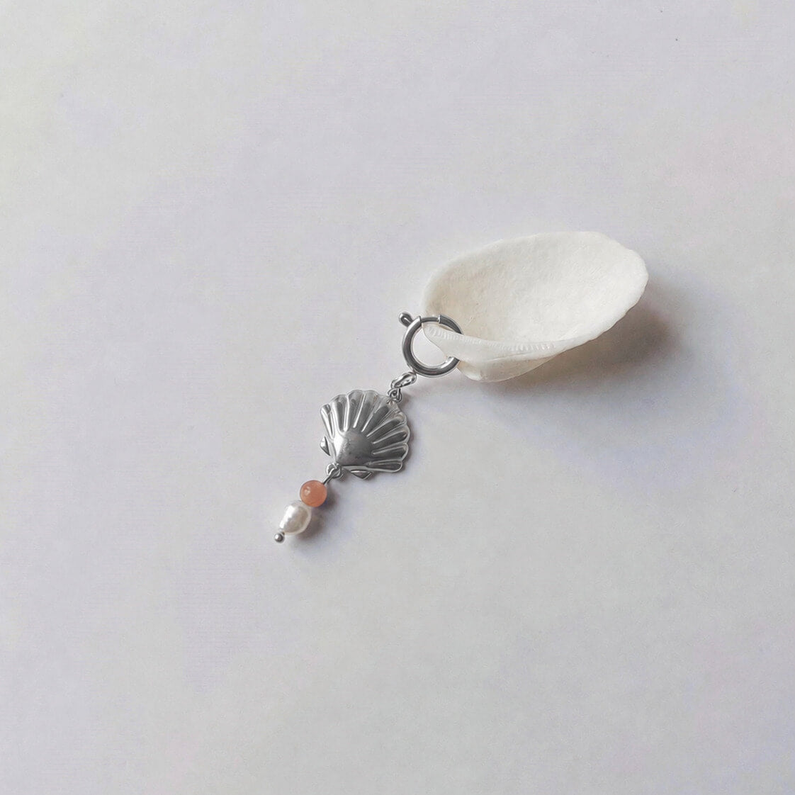 Charm à anneau ressort argenté composé d'un pendentif coquille Saint-Jacques argentée striée, d'une petite perle de pierre de soleil et d'une perle d'eau douce blanche baroque, posé contre un coquillage nacré blanc sur fond gris clair.