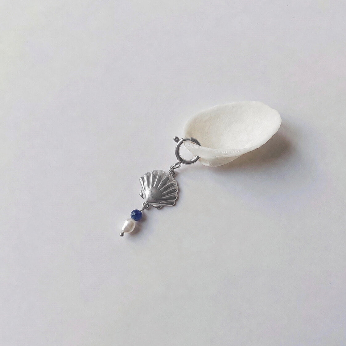 Charm à anneau ressort argenté composé d'un pendentif coquille Saint-Jacques argentée striée, d'une petite perle de sodalite et d'une perle d'eau douce blanche baroque, posé contre un coquillage nacré blanc sur fond gris clair.