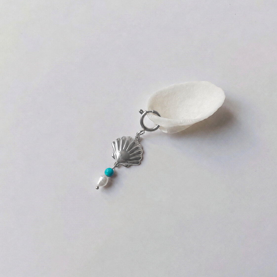 Charm à anneau ressort argenté composé d'un pendentif coquille Saint-Jacques argentée striée, d'une petite perle de turquoise et d'une perle d'eau douce blanche baroque, posé contre un coquillage nacré blanc sur fond gris clair.