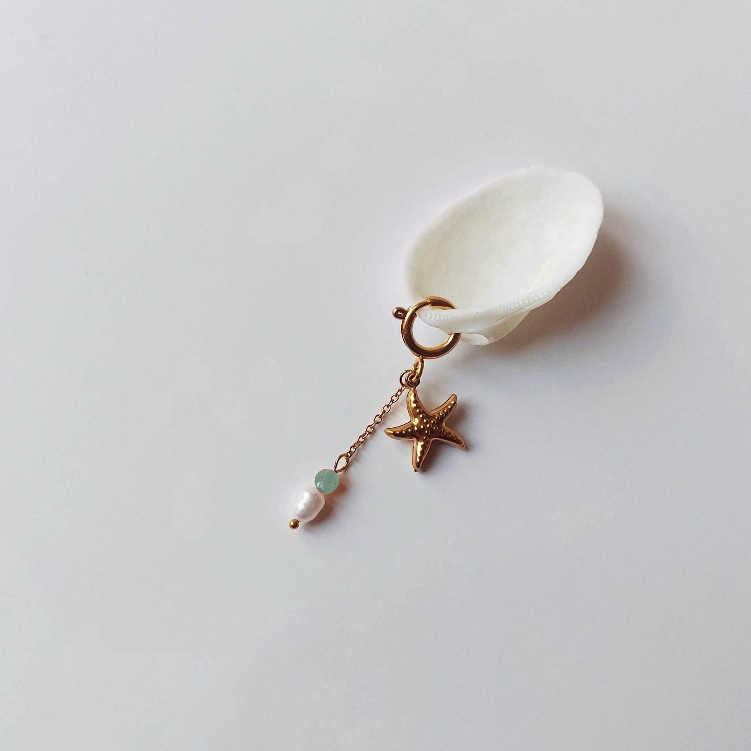 Charm à anneau ressort doré composé d'un pendentif étoile de mer dorée texturée et d'une chaînette fine ornée d'une petite perle d'aventurine et d'une perle d'eau douce blanche baroque, posé contre un coquillage nacré blanc sur fond gris clair.