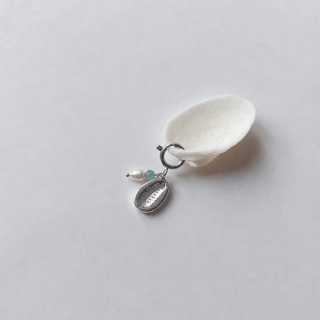 Charm à anneau ressort doré composé d'une perle d'eau douce blanche, d'une petite perle d'aventurine et d'un pendentif cauri doré, posé contre un coquillage nacré blanc sur fond gris clair.