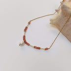 Collier Anthozoa en pierres de quartz fraise roses et petites perles d'eau douce encadrées de rondelles dorées fleuries, avec une perle d'eau douce baroque en pendentif central, sur fine chaîne dorée en acier inoxydable, posé contre un grand coquillage beige sur fond blanc.
