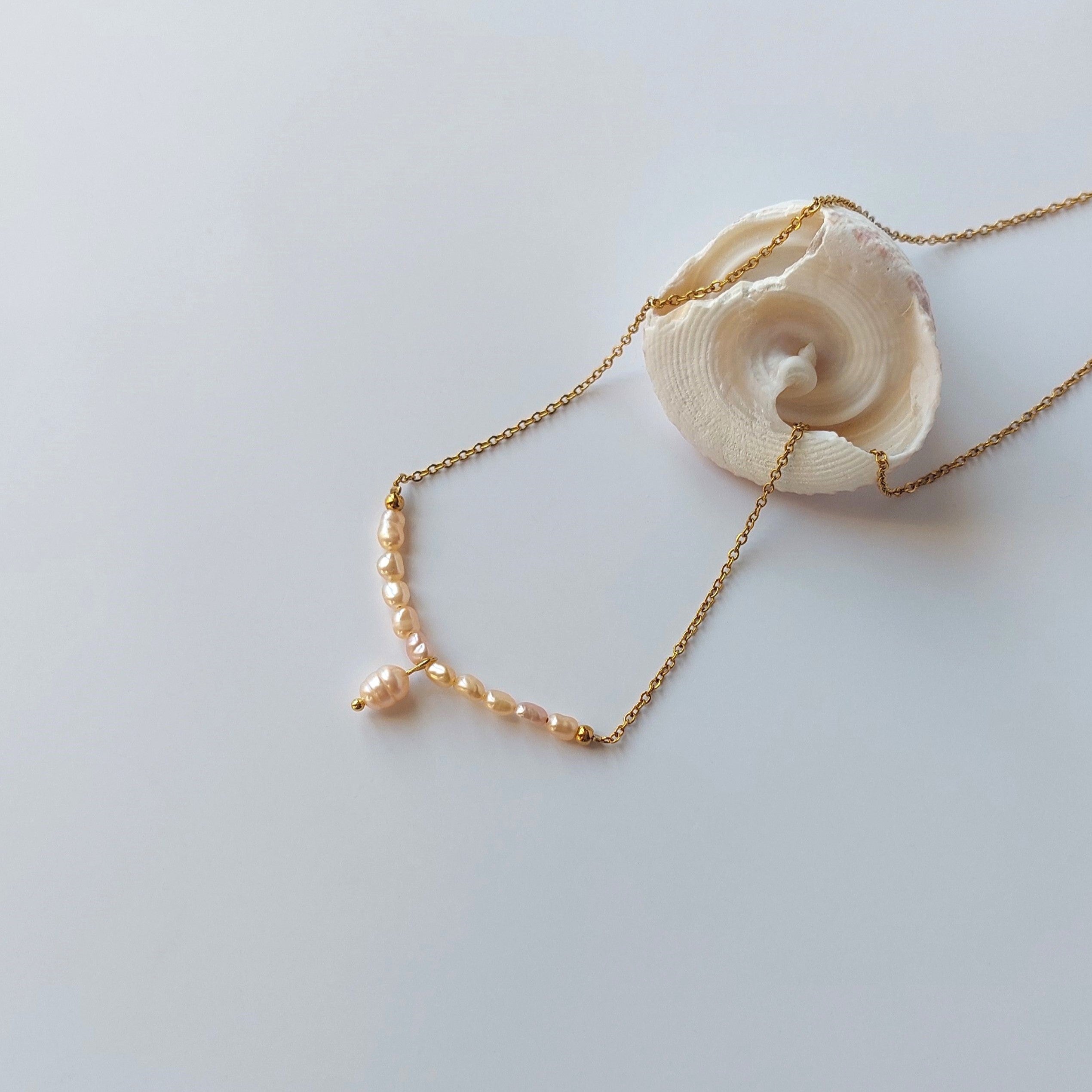 Collier Aurea en acier inoxydable doré avec perles d’eau douce naturelles oranges, bijou fin et élégant posé sur un coquillage, sur fond neutre.