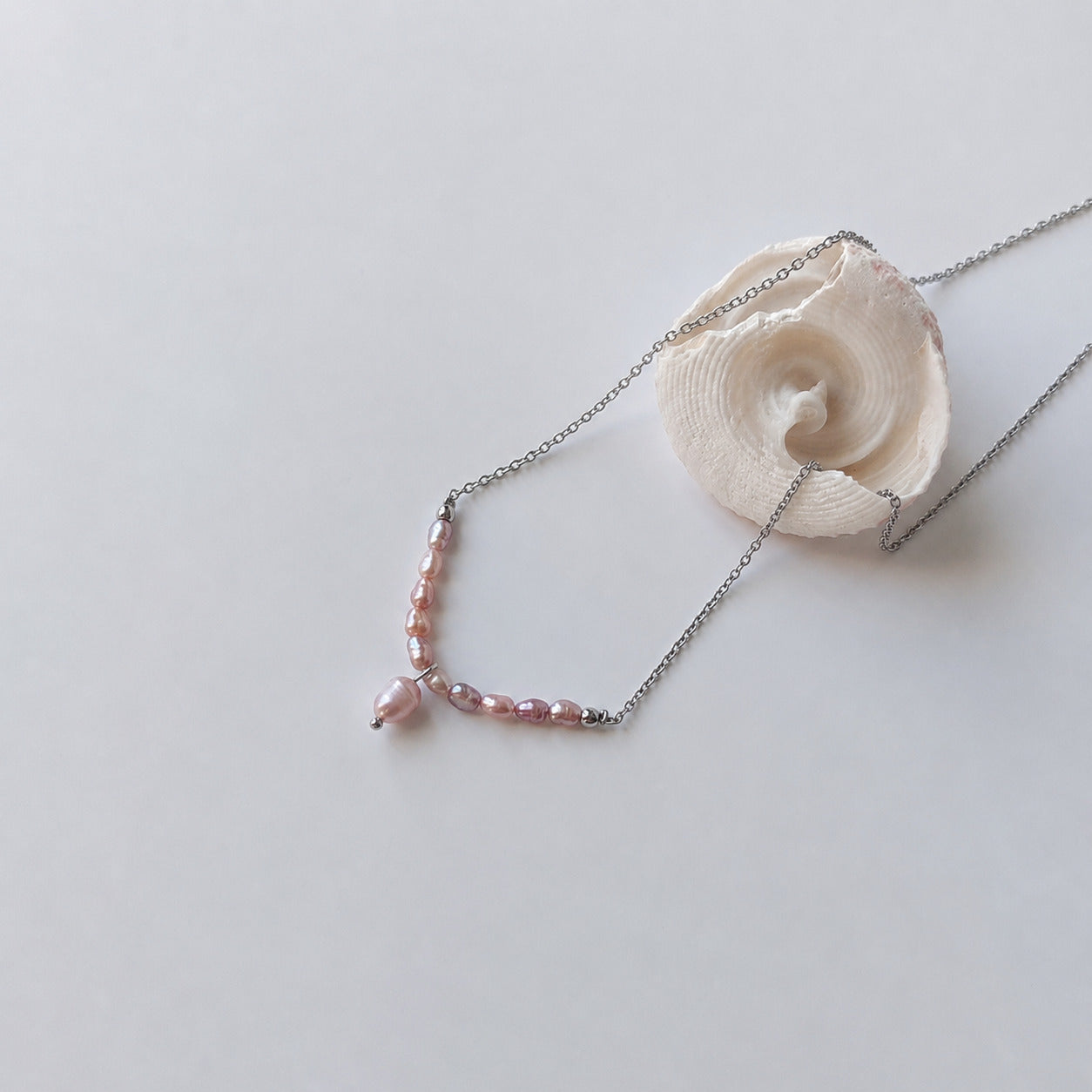 Collier fin sur chaîne argentée composé d'une rangée de petites perles d'eau douce violettes baroques en arc, avec une perle plus grande en pendentif central, posé sur fond gris clair aux côtés d'un coquillage en spirale blanc.