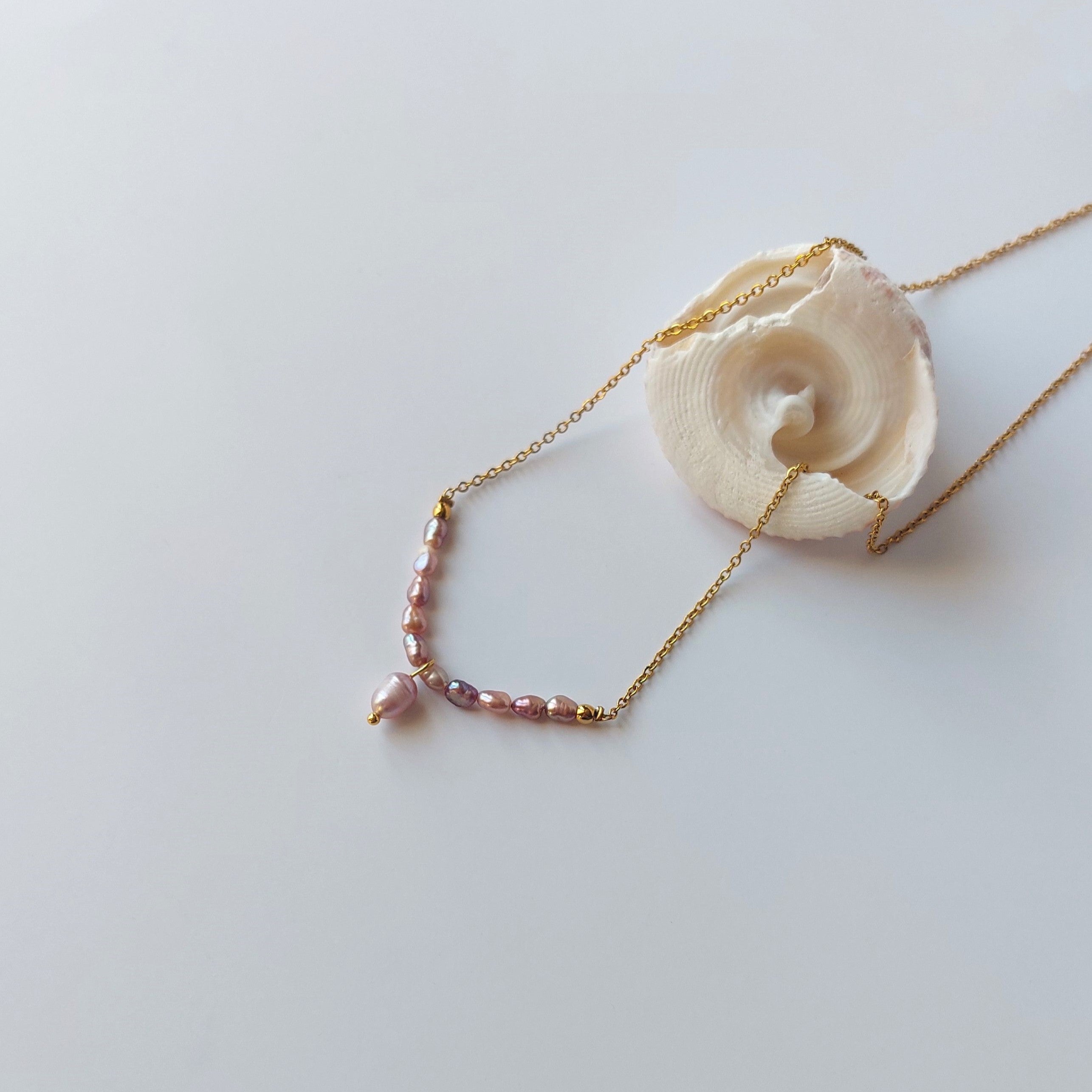 Collier Aurea en acier inoxydable doré avec perles d’eau douce naturelles violettes, bijou fin et élégant posé sur un coquillage, sur fond neutre.